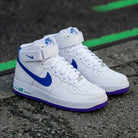 A pair of Nike Air Force 1 Mid sneakers in white, featuring a vibrant blue Swoosh and a purple outsole, resting on an asphalt surface with green painted lines.